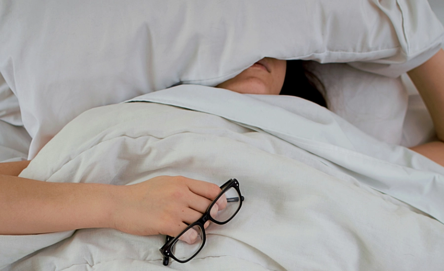 Person lying on bed while covering face with pillow and holding eyeglasses.