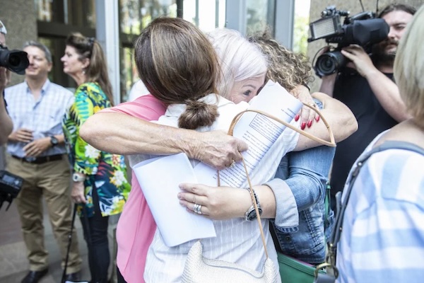 Three women hug outside a courthouse, surrounded by cameras