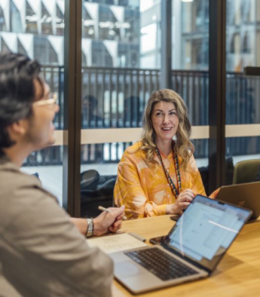 Two uni staff members with laptops sit at converence table in conversation.