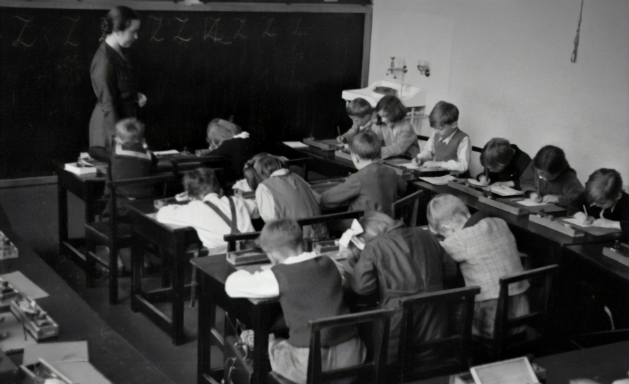 Grayscale photography of teacher standing near chalkboard and children sitting on chairs.
